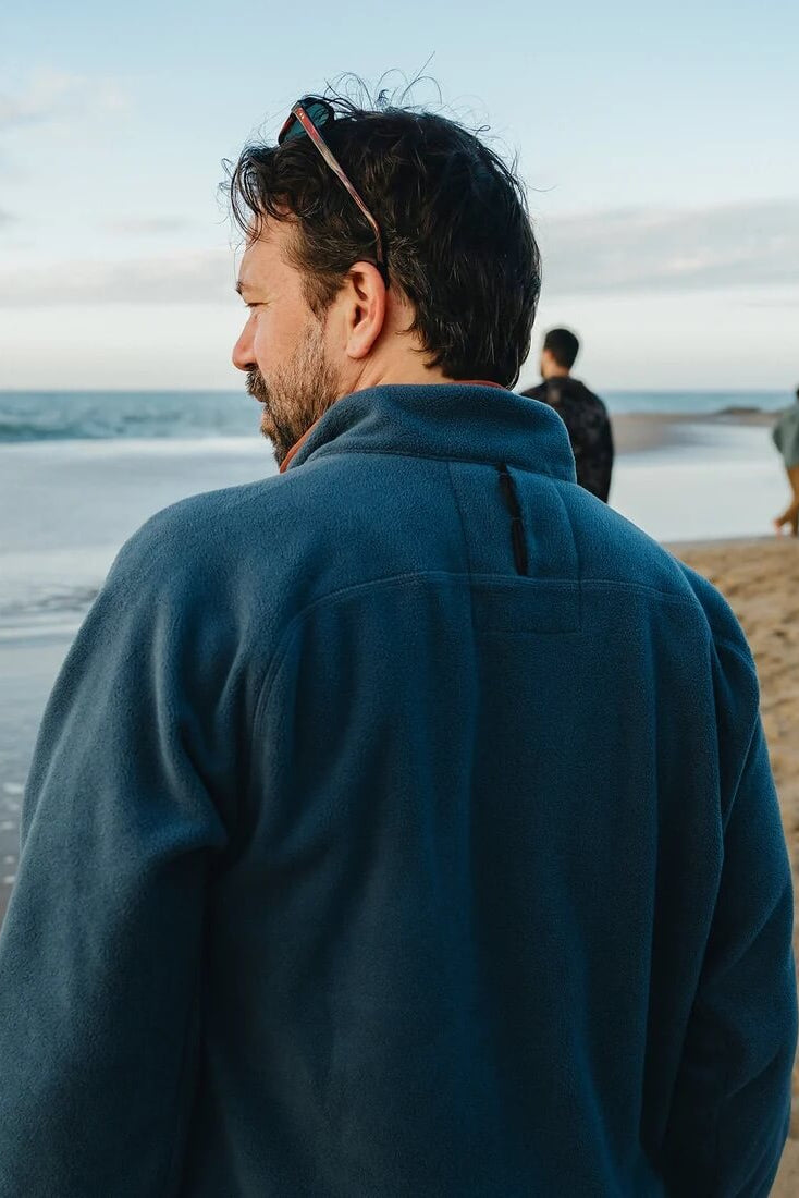 A man wearing Howler Bros Free Range Fleece Pullover in Nightfall, seen from behind as he looks out toward the water.