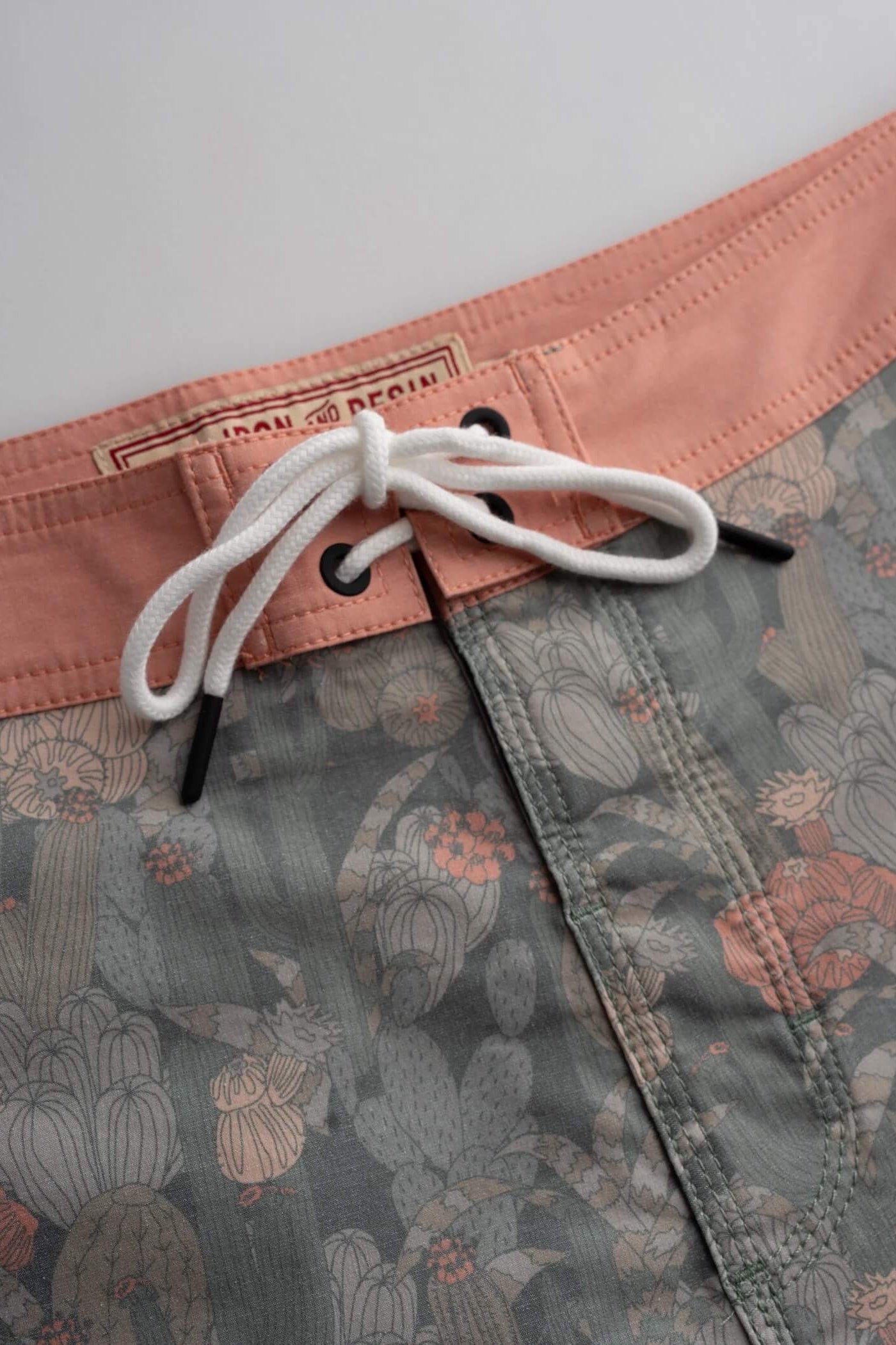 Close-up of board shorts showing a peach waistband, white drawstring, and a muted gray-green floral and cactus print fabric.