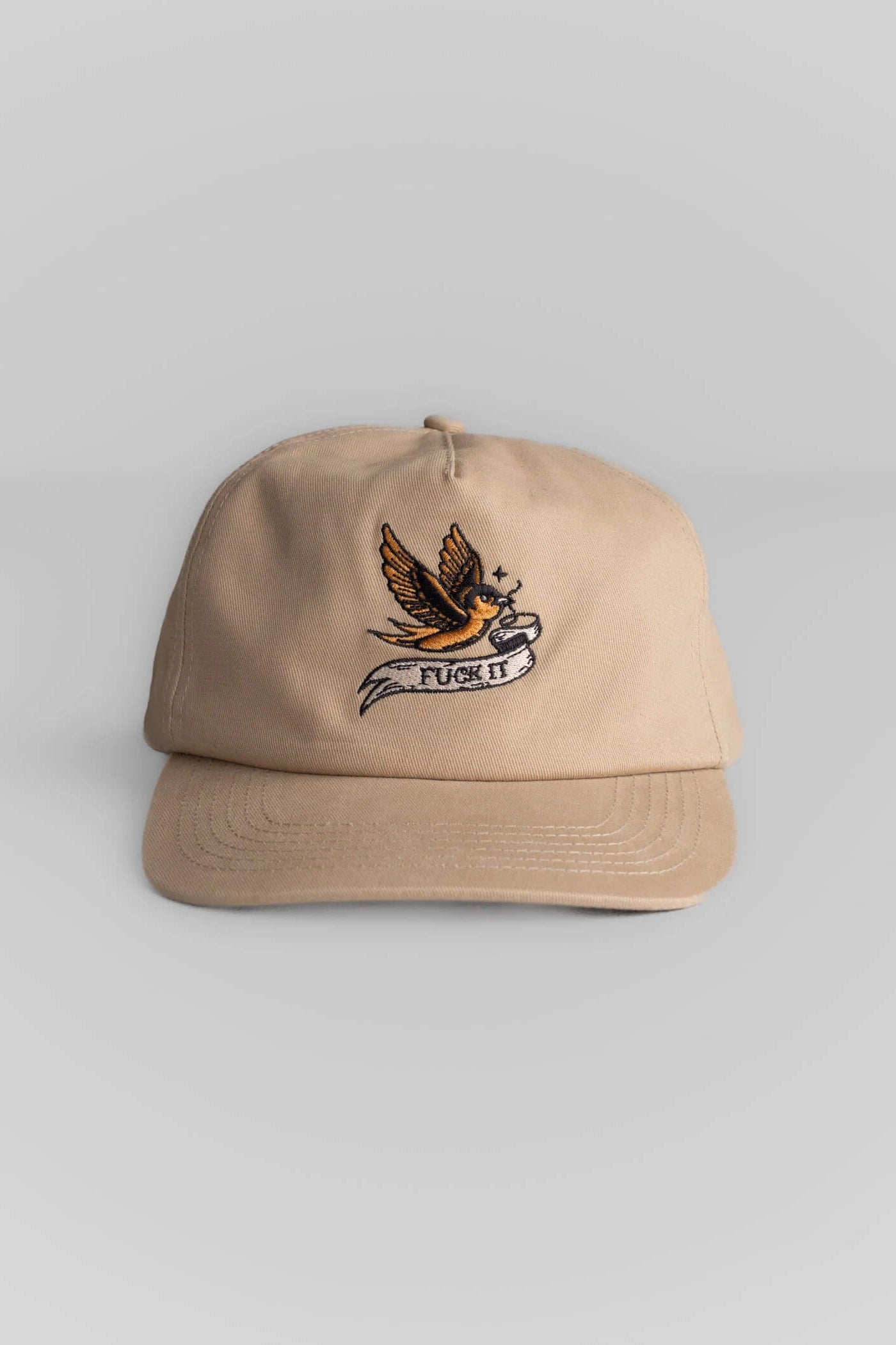 A beige baseball cap featuring an embroidered graphic of a bird holding a banner that reads 'PUSH IT' in orange, black, and white.