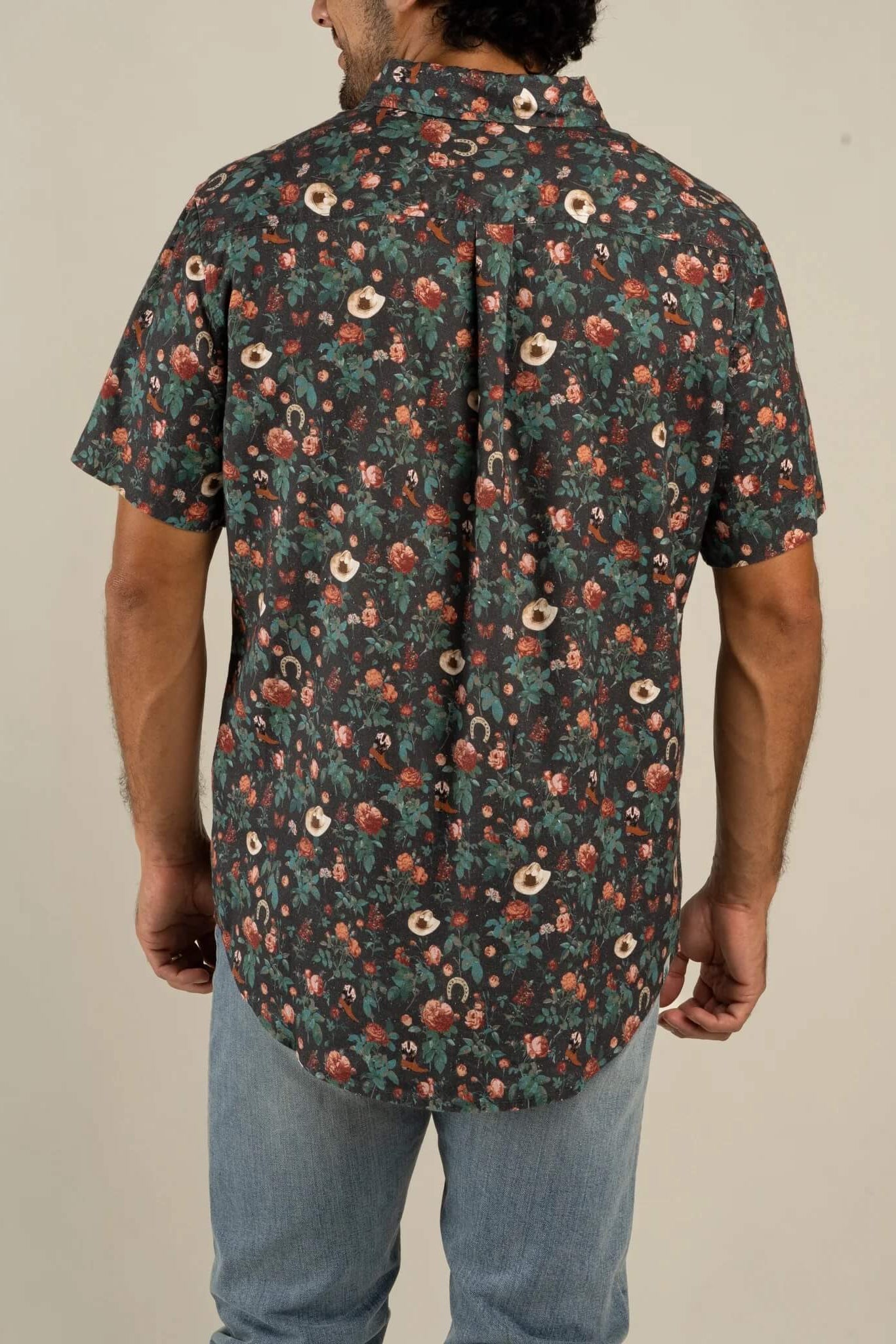 A man viewed from the back wearing a short-sleeve button-up shirt with an all-over dense floral print in dark green, rust red, and cream, paired with light blue jeans against a plain beige studio background.