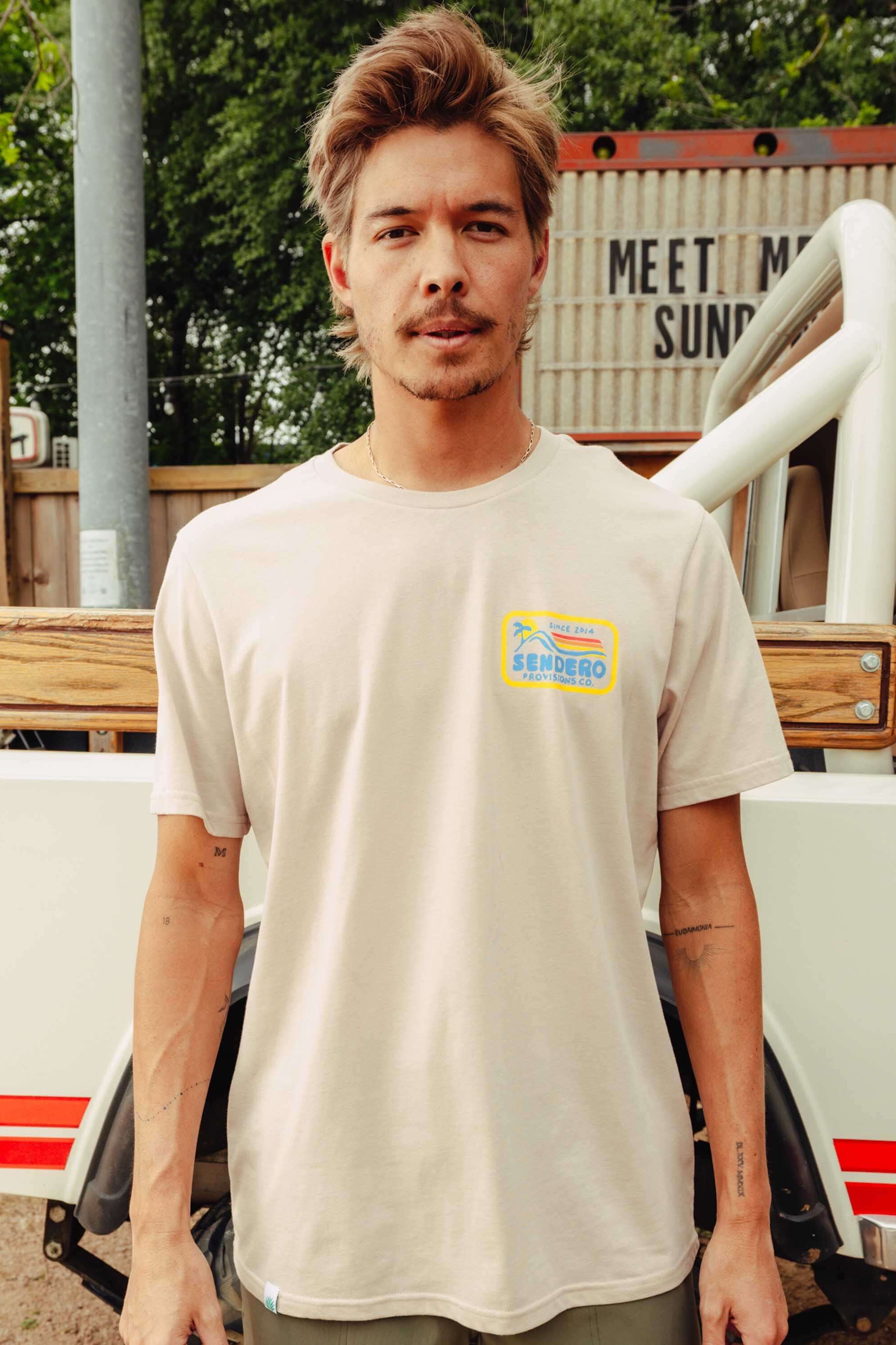A man wearing a light beige crewneck t-shirt with a small yellow and blue graphic on the left chest, standing outdoors in front of a white and red vehicle.
