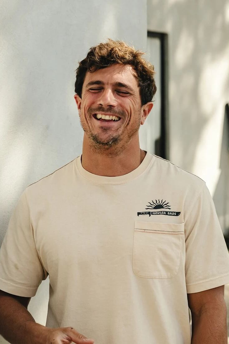 A smiling man wearing Howler Bros HB Machete Cotton T-Shirt in Sand, standing against a white stucco wall.