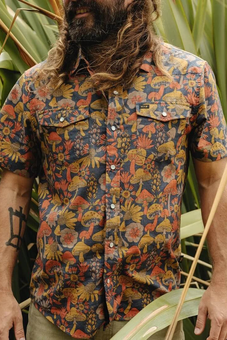 A man wearing Howler Bros H Bar B Snapshirt in Fungi Forage- Nightfall, standing in front of green tropical leaves.