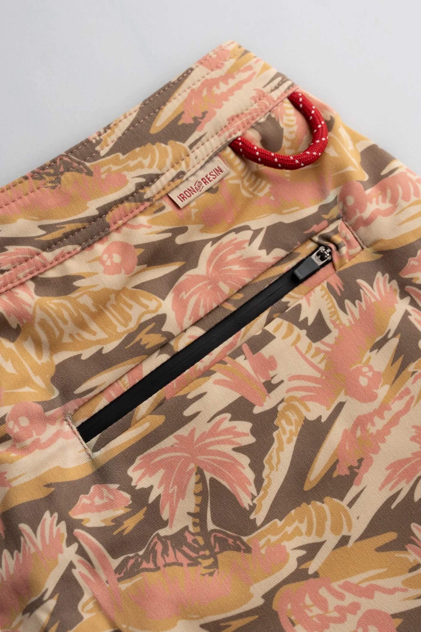 Close-up of a pair of shorts featuring a tropical palm tree print in beige, pink, and brown, with a black zipper pocket and a red beaded ring detail near the waistband.
