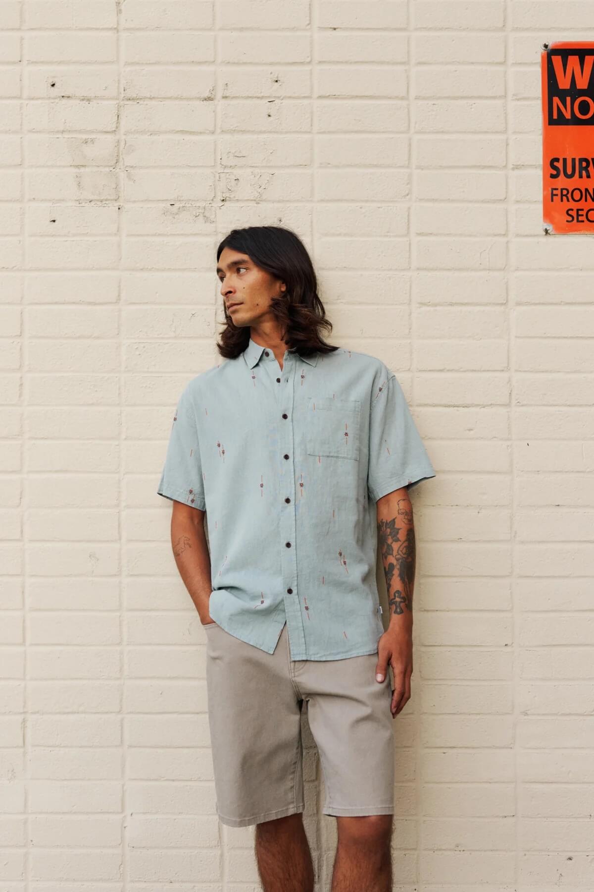 Man wearing a light blue shirt and Katin George 64 Bull Denim Short  standing against a white brick wall.