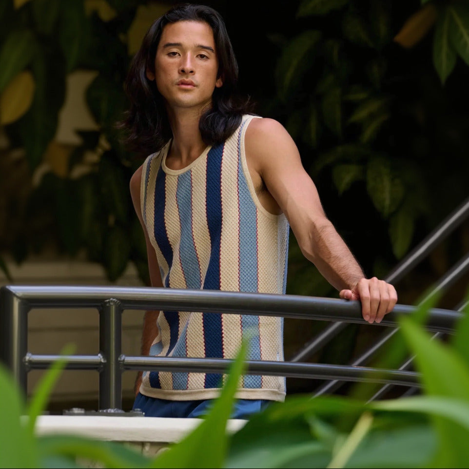 Man wearing a sleeveless striped shirt standing outdoors with plants in the background