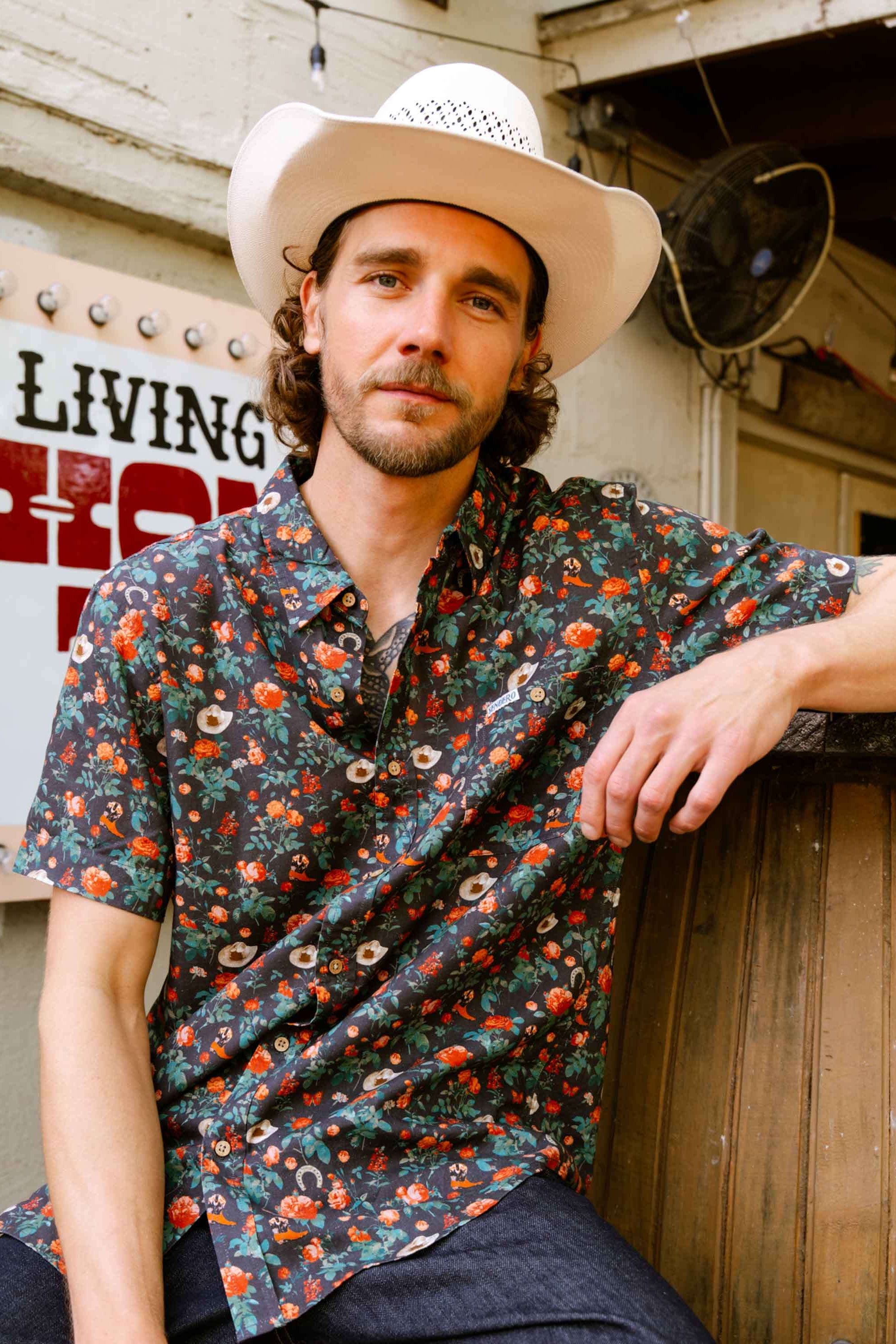 Man wearing a cowboy hat and City Slicker shirt leaning against a wooden structure 