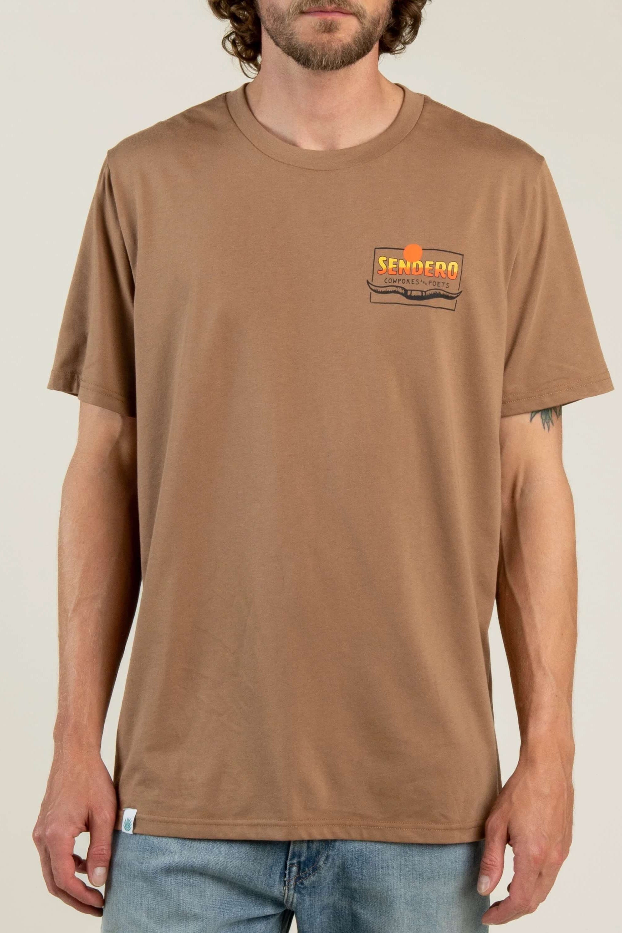 A man wearing a brown crewneck t-shirt with a small 'Kemper' logo patch on the left chest, paired with blue jeans, against a light beige background.