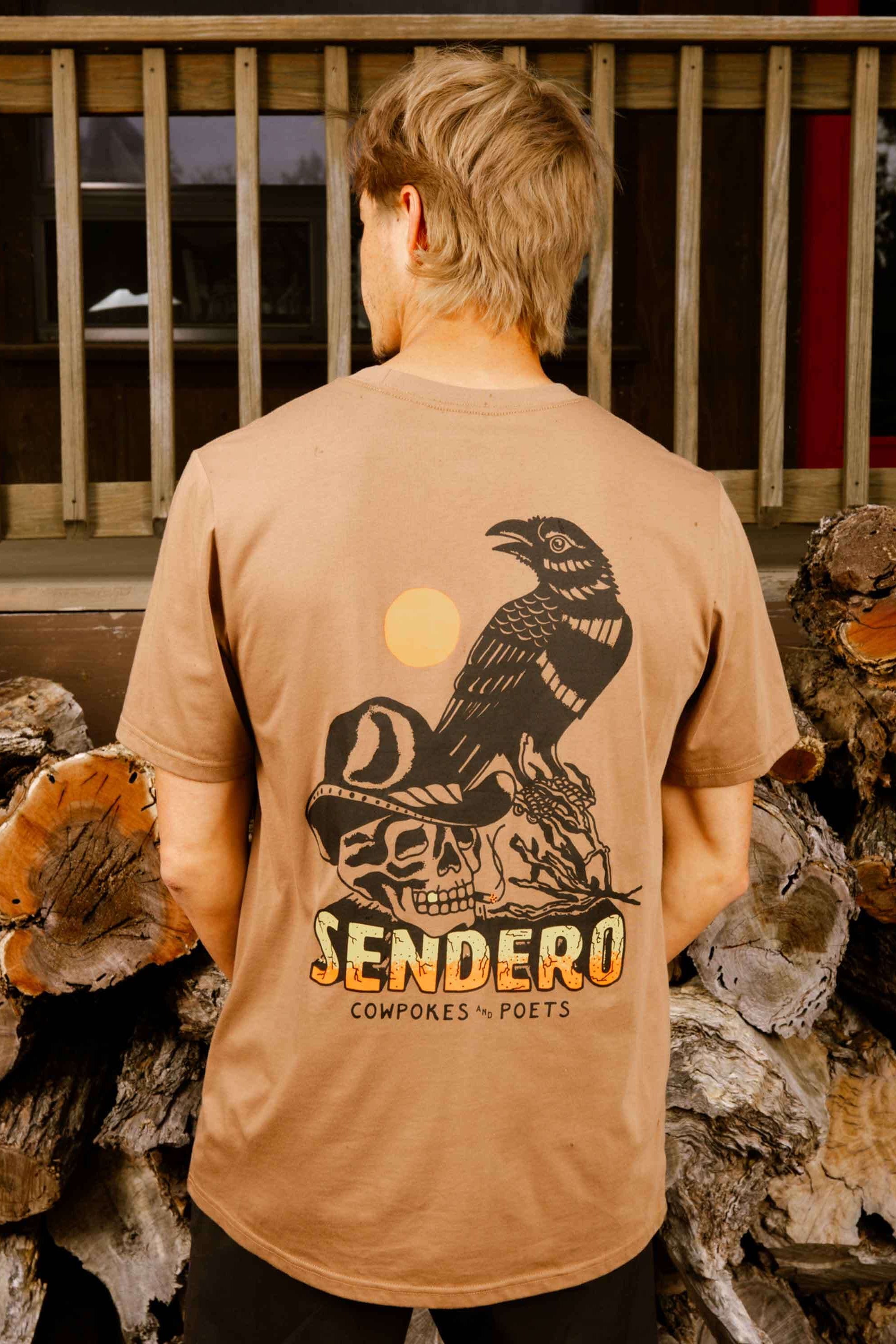 Person wearing a beige t-shirt with a graphic design and 'Sendero' text, standing in front of stacked logs.